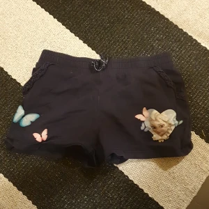 Svarta Disney shorts med fjärilar från H&M - Svarta shorts från H&M med Disney-tryck, featuring Elsa från Frost i ett hjärta och färgglada fjärilar på framsidan. Mjuk bomull, elastisk midja med snörning och fickor med flätad detalj. Perfekta för chill dagar och sommarhäng.