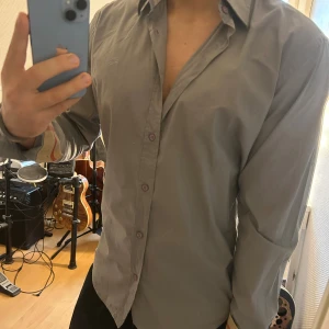 Grå Burberry Brit skjorta L - Snygg grå skjorta från Burberry Brit i storlek L. Klassisk modell med knappar framtill och långärmade ärmar. Insidan av manschetterna har det ikoniska rutiga Burberry-mönstret i beige, blått och rött. Skjortan är gjord i mjuk bomull och har en normal passform.