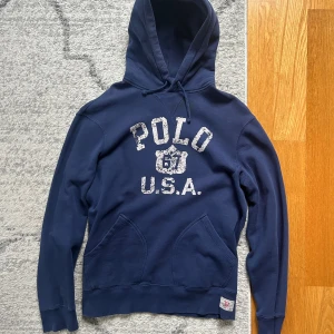 Marinblå hoodie från Polo Ralph Lauren - Snygg marinblå hoodie från Polo Ralph Lauren med vit vintageinspirerad logga 'POLO U.S.A.' framtill. Tröjan har huva med snörning och en stor magficka. Perfekt för en chill och sportig look.
