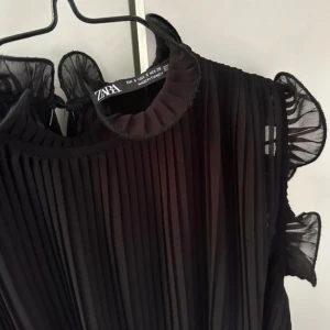 Svart plisserad topp från Zara - Svart plisserad topp från Zara med transparenta volangdetaljer vid ärmhål och halsringning. Toppen  är ärmlös och har en luftig, loose passform som ger en cool och elegant vibe. Perfekt för dig som gillar stilrena och trendiga plagg.