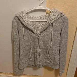 Grå hoodie med teddyfoder från Hollister - Mysig grå hoodie från Hollister med broderad logga och årtal på bröstet. Tröjan har dragkedja, två fickor fram, snörning i huvan och är fodrad med fluffigt teddyfoder för extra värme. Perfekt för kyliga dagar. Är storlek S man är liten i storlek kanske passar dig som bär XS