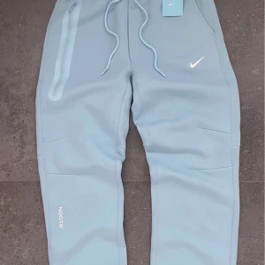 Nike NOCTA ljusblå mjukisbyxor S - Nike NOCTA mjukisbyxor i ljusblå färg med vit logga och snörning i midjan. Byxorna har dragkedjeficka på sidan, elastisk midja och tryckta detaljer som NOCTA, C.L. och A.W.R. Skön passform och stilren design, perfekt för chill eller streetwear.