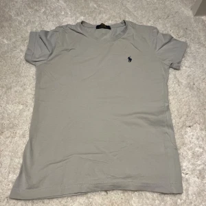 Grå t-shirt från Polo Ralph Lauren - Snygg grå t-shirt från Polo Ralph Lauren i mjuk bomull. Klassisk passform med korta ärmar och rund hals. Diskret mörkblå broderad logga på bröstet ger en clean och stilren look. Perfekt för dig som gillar enkel och tidlös design.