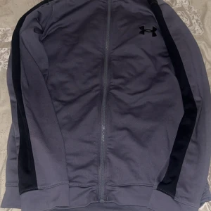 Grå träningsjacka från Under Armour - Snygg grå träningsjacka från Under Armour med svarta detaljer längs ärmarna och dragkedja framtill. Jackan har hög krage och två sidofickor, perfekt för att hålla stilen på gymmet eller ute. Loggan syns på bröstet för extra sportig vibe.