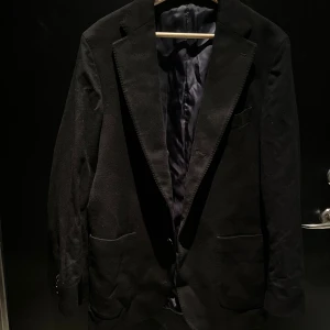 Jacka Rose & Born - Stilren svart kavaj/jacka från Rose & Born, tillverkad i exklusivt tyg från Loro Piana. Klassisk modell med två framfickor, bröstficka och knappar vid ärmslut. Fodrad insida i mörk nyans. Perfekt för dig som vill ha en tidlös och elegant look. Ordinariepris: runt 18.000kr