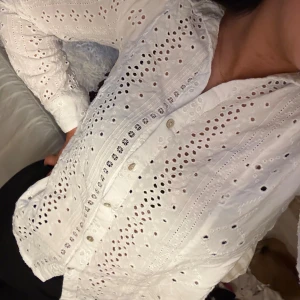 Vit broderad skjorta med spetsdetaljer - Säljer en vit skjorta med broderade spetsdetaljer och hålmönster. Skjortan har knappar framtill, lång ärm och en snygg v-ringad krage. Materialet är mjukt och känns som bomull, perfekt för dig som gillar romantisk och chill stil.