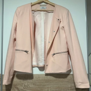 Ljusrosa kavaj från ONLY - Snygg ljusrosa kavaj från ONLY med stilrena dragkedjor vid fickorna och rundad halsringning. Jackan har lång ärm och är lätt figursydd för en modern look. Perfekt att slänga över en t-shirt eller topp för att lyfta din outfit.