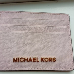 Rosa plånbok från Michael Kors - Snygg och stilren rosa plånbok från Michael Kors med flera kortfack och ett fönster för ID eller busskort. Plånboken har guldfärgad logotyp framtill och är tillverkad i ett strukturerat syntetmaterial som känns lyxigt och modernt.