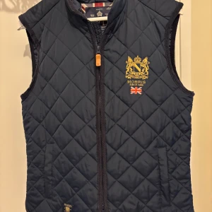 Mörkblå quiltad dunväst från Morris - Nästan aldrig änvänd så är ny skick. Quiltad mörkblå dunväst från Morris med broderad gul logga och brittisk flagga på bröstet. Västen har dragkedja framtill, ståkrage och två fickor. Perfekt för lager-på-lager och snygg till både hoodie och skjorta. perfekt om du är 165-175 då den är lite större i storlek 