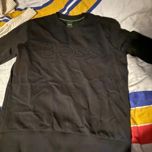 Svart sweatshirt från BOSS - Svart sweatshirt från BOSS med broderad logga framtill. Tröjan har rund hals och långa ärmar, samt ribbade muddar vid ärmslut och nederkant. Perfekt för dig som gillar stilrena och klassiska plagg med snygg branding.