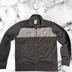 Nike Air svart och grå zip - Nike Air zip i svart och grått med blå swoosh på bröstet. Jackan har hel dragkedja, ståkrage och två sidofickor. Materialet är mjukt och syntetiskt, perfekt för en sportig look. AIR-tryck på framsidan ger extra stilpoäng.