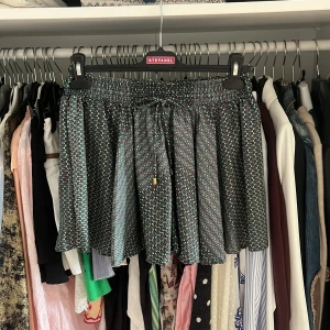 Volang kjol från copenhagen luxury - Snygg kjol med grönt och svart småmönster, elastisk midja och snörning framtill med gulddetaljer. Kort passform, perfekta för varma dagar. Tunt, glansigt material som känns lyxigt och bekvämt.