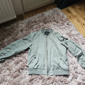 Ljusgrön bomberjacka från Zoul Edition - Snygg ljusgrön bomberjacka från Zoul Edition i storlek M. Jackan har dragkedja framtill, fickor med tryckknappar och en extra ficka med dragkedja på ärmen. Tillverkad i polyester och polyamid, med ribbade muddar och klassisk bomberkrage.