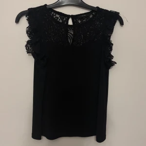 Svart spetsblus från Vero Moda - Supersnygg svart blus från Vero Moda med spets upptill och volangdetaljer vid axlarna. Ärmlös modell med rund halsringning och knapp i nacken. Perfekt för dig som vill ha en stilren men ändå detaljrik topp.