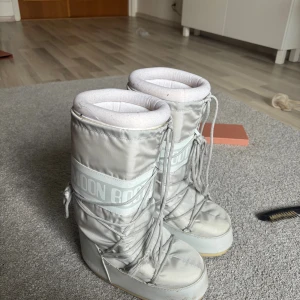 Grå Moon Boots vinterstövlar - Säljer ett par klassiska grå Moon Boots med hög skaft och snörning framtill. De är tillverkade i syntetmaterial med vadderad insida och bred sula, perfekta för kalla vinterdagar. Stilen är ikonisk och ger en cool retrovibe till din vinteroutfit.
