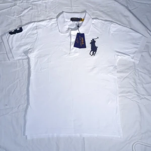 Vit pikétröja från Polo Ralph Lauren - Vit pikétröja från Polo Ralph Lauren med klassisk krage, knappar och stor mörkblå broderad logga på bröstet. Slim fit-modell med korta ärmar och siffra på ärmen. Perfekt för en clean och sportig stil.