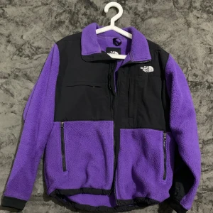Lila fleecejacka från The North Face - Säljer en lila och svart fleecejacka från The North Face med dragkedja framtill, höga fickor och broderad logga på bröstet. Jackan har hög krage och lång ärm, perfekt för kyliga dagar. Snygg och sportig stil som sticker ut.