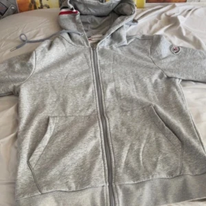 Grå Moncler hoodie med dragkedja - Snygg grå hoodie från Moncler med hel dragkedja, huva med snörning och röd, vit och marinblå randdetalj. Tröjan har två fickor framtill och Moncler-logga på ärmen. Perfekt för chill dagar och streetstyle.