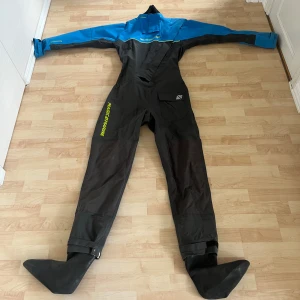 Magic Marine blå och svart torrdräkt för segling - Magic Marine torrdräkt i blått och svart med gul logga och detaljer. Jackan har hög krage, långa ärmar med justerbara muddar och dragkedja framtill. Tillverkad i slitstark syntet, perfekt för vattenaktiviteter. Snygg och funktionell design med praktisk ficka på benet. Inga hål, nyköpt och använd under två säsonger. Kostade orginellt 7000kr.
