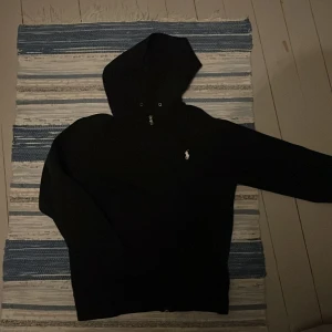 Svart hoodie från Ralph Lauren - Svart hoodie från Ralph Lauren med klassisk vit logga på bröstet. Tröjan har huva med snörning och långa ärmar. Perfekt för en chill och stilren look.