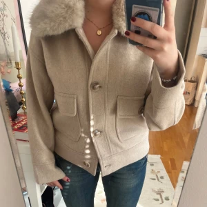 Beige ulljacka Neo Noir - Snygg beige jacka från Neo Noir med fluffig päls på kragen och rejäla fickor framtill. Jackan har knappar hela vägen och är tillverkad i mjuk ullmix med fodrad insida. Perfekt höstjacka. Jackan är i nyskick förutom en rem på pälsen som lossnat (sista bilden) men inget som påverkar då den sitter fast med knappar. Nypris 1900