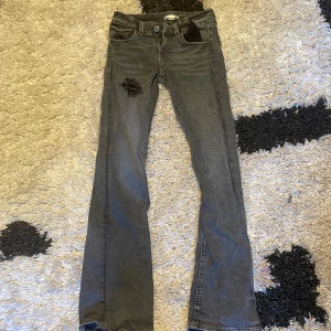 Grå bootcut jeans med detaljer - Säljer ett par grå jeans med bootcut passform och snygga slitningar framtill. Baksidan har broderade fickor med dekorativa sömmar och knappar. Jeansen har låg midja och är tillverkade i klassiskt denimtyg. Perfekta för dig som gillar en lite mer edgy stil.