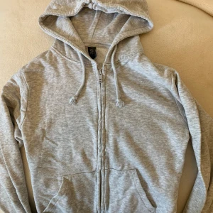 Grå hoodie med dragkedja från SOC XS - Mjuk och skön ljusgrå hoodie från SOC i storlek XS. Tröjan har huva med snörning, dragkedja framtill och två fickor. Perfekt för chill dagar eller till träningen.