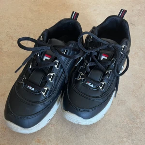 Svarta Fila sneakers chunky sula - Chunky svarta Fila sneakers med vit, kraftig sula och klassisk logga på sidan och plösen. Skorna har snörning med metallöglor och är tillverkade i syntetmaterial. Perfekta för dig som gillar streetstyle och vill ha en sportig vibe.