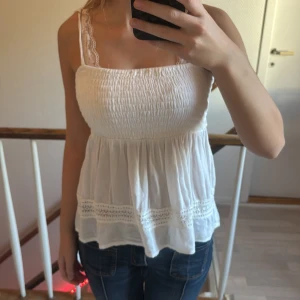 Vit smockad linnetopp med spets - Söt vit linnetopp med smockad överdel och smala axelband med spetsdetaljer. Toppen har en luftig, volangig nederdel och dekorativ spets längs fållen. Perfekt för varma dagar och enkel att matcha med jeans eller kjol.