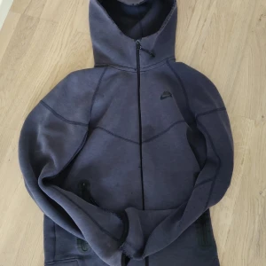 Mörkblå Nike tech  hoodie med dragkedja - Nike hoodie i mörkblått med hel dragkedja och huva. Tröjan har svarta detaljer, två fickor med dragkedja och diskret Nike-logga på bröstet. Perfekt för en sportig och avslappnad stil.