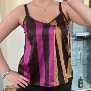 Randig linnetopp från Vero Moda - Säljer en snygg linnetopp från Vero Moda med breda vertikala ränder i brunt, lila och beige. Toppen har smala axelband och v-ringning framtill. Perfekt för dig som gillar färg och mönster med en cool retrovibe. Axelbredd: 27cm. Längd: 60cm. Byst: 40cm.