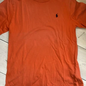 Orange t-shirt från Ralph Lauren, strl 10-12 - Snygg orange t-shirt från Ralph Lauren med klassisk mörkblå logga broderad på bröstet. T-shirten har rund halsringning och korta ärmar, perfekt för en avslappnad och stilren look.