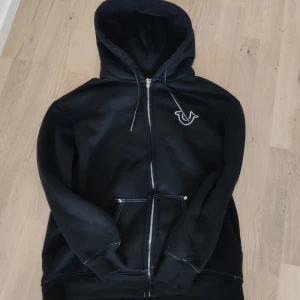  True Religion hoodie  - Svart hoodie från True Religion med vit logga på bröstet och stort tryck på ryggen. Dragkedja framtill, huva med snörning och känguruficka. Snygga kontrastsömmar och klassisk streetwear-vibe. Perfekt för dig som gillar stilrena och coola plagg.