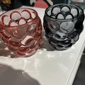 Ljus behållare - Bilderna visar två dekorativa glas med bubblig struktur, ett i rött och ett i grått. De har en unik design och ser ut att vara i mycket gott skick utan synliga defekter eller slitage. Perfekta för servering eller som inredningsdetalj.