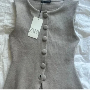 Beige stickad väst från Zara - Supersnygg beige stickad väst från Zara med stora klädda knappar framtill. Västen är ärmlös, har en rak passform och en liten slits nertill. Perfekt att styla över en skjorta eller t-shirt för en trendig look.