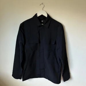 Svart overshirt från H&M, strl 48 - Stilren svart overshirt från H&M med klassisk krage och två bröstfickor med lock. Skjortan är tillverkad i en mix av bomull och polyamid, har långa ärmar med manschett och knappar framtill. Perfekt för lager-på-lager och enkel att matcha. 3U4
