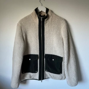 Beige teddyjacka från Selected Homme - Mysig beige teddyjacka från Selected Homme med svarta detaljer och stora fickor framtill. Jackan har hög krage, hel dragkedja och är tillverkad i 100% polyester. Perfekt för kalla dagar när du vill ha både stil och komfort. 3W7