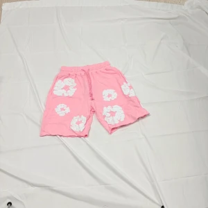 Rosa shorts från Denim Tears med blommor - Säljer ett par rosa shorts från Denim Tears med vita blommiga mönster. De har elastisk midja med snörning och är tillverkade i mjuk bomull. Perfekta för en chill sommarlook med sin loose fit och unika design.