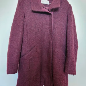 Vinröd ullkappa från Esprit, stl 36 - Vinröd kappa från Esprit i storlek 36. Kappan är tillverkad i ullblandning med en mysig, lite ruggad yta och har klassisk krage samt två snedställda fickor framtill. Perfekt för kyliga dagar och ger en stilren vibe till din outfit.