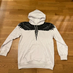 Vit hoodie Marcelo Burlon med svarta vingar - Säljer en vit hoodie från Marcelo Burlon County of Milan med ikoniskt svart vingtryck över axlar och bröst. Tröjan har huva med snörning, känguruficka och långa ärmar. Tillverkad i mjuk bomull, perfekt för dig som vill sticka ut med en unik design.