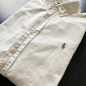 Vit skjorta från Polo Ralph Lauren - Klassisk vit skjorta från Polo Ralph Lauren i storlek L, custom fit. Skjortan har knappar framtill och den ikoniska broderade polospelaren på bröstet. Tillverkad i mjuk bomull med krage och stilren design, perfekt för dig som gillar clean och tidlös stil.