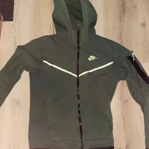 Grön Nike hoodie med dragkedja - Snygg grön hoodie från Nike med hel dragkedja och huva. Tröjan har en vit Nike-logga på bröstet, vita detaljer över bröstet och en praktisk ficka med dragkedja på ena ärmen. Perfekt för chill eller träning.