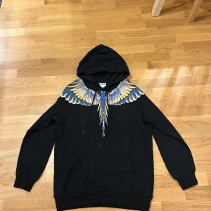 Svart hoodie Marcelo Burlon med vingar - Svart hoodie från Marcelo Burlon County of Milan med ikoniskt vingtryck i blått och gult över axlar och bröst. Tröjan har huva med snörning och är tillverkad i mjuk bomull. Perfekt statement-plagg för dig som vill sticka ut.