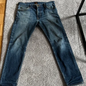 Blå Lee Rider jeans W31 L32 - Säljer ett par klassiska blå Lee Rider jeans i rak modell. Jeansen har snygga slitningar och kontrastsömmar, fem fickor och Lee-logga på knappen. Tillverkade i bomull för en skön känsla och passform. Perfekta för dig som gillar tidlös denimstil. En litt hål se på bilden! 