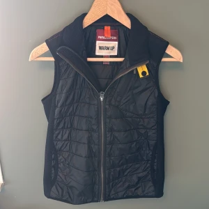 Svart dunväst från Parajumpers - Svart quiltad dunväst från Parajumpers med dragkedja framtill, hög krage och ikonisk gul detalj vid halsen. Västen har sportig look, sidfickor och diskret logga i nacken. Perfekt för lager-på-lager och outdoor-vibe.