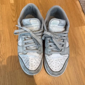 Nike Dunk sneakers i ljusgrå mocka - Nike Dunk sneakers i ljusgrå mocka och vitt, med klassisk snörning och perforerad tå. Snygg och tidlös modell som passar perfekt till streetwear och vardagsstil. Diskret Nike-logga på tungan och sidan. Bekväm platt sula och rund tå.