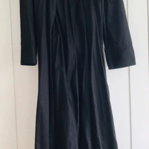 Svart långklänning från Zara - Svart långklänning från Zara med knappar hela vägen framtill. Klänningen har trekvartsärmar och en rak, avslappnad passform. Tillverkad i ett lätt, glansigt material som ger en stilren look. Perfekt för dig som gillar minimalistisk stil. Köpt för 500 kr men du får det till ett billigt pris!