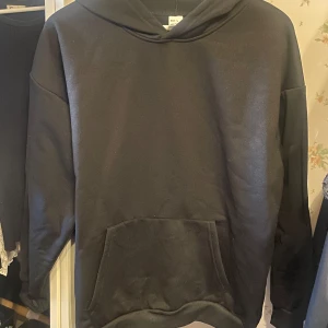 Svart hoodie i polyester, storlek L - En svart hoodie i storlek L med klassisk känguruficka framtill och huva. Tillverkad i 100% polyester, perfekt för dig som gillar en enkel och stilren look. Passformen är normal och modellen har långa ärmar.
