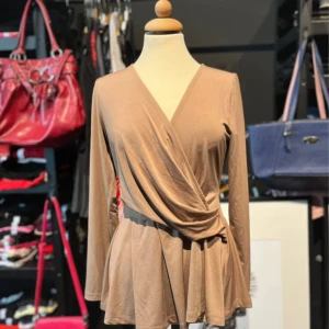 Beige omlottopp  - Säljer en beige omlottopp från Chiara For Milano i mjuk och stretchig polyesterblandning. Toppen har lång ärm, v-ringning och ett snyggt omlottfall som ger en avslappnad look. Perfekt att styla med jeans