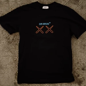 Off-White svart t-shirt med KAWS print - Svart t-shirt från Off-White med färgstarkt KAWS-inspirerat print på ryggen och Off-White-logga med två orange kryss på bröstet. T-shirten är i bomull och har en klassisk passform med korta ärmar. Perfekt för dig som gillar streetwear och unika designer.